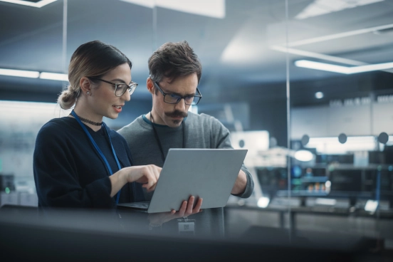 A photograph of a man and a woman standing side by side looking at a laptop screen in an office environment