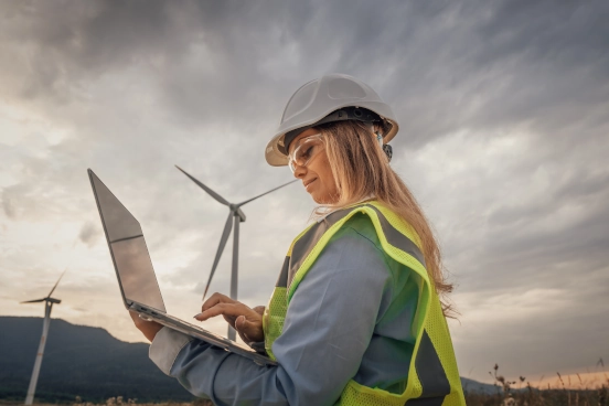 A photo of a person wearing a high vis top and a hard hat in front of a wind turbine operating a laptop computer