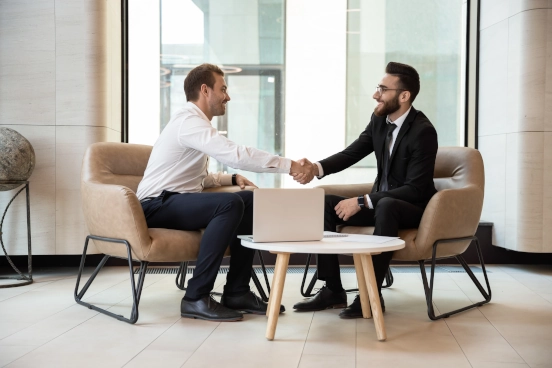 A photo of two men smiling in business formal attire sat at a table reaching across to shake hands
