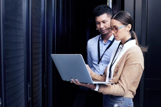 A photograph of two people stood in a datacentre discussing the content of a laptop screen.